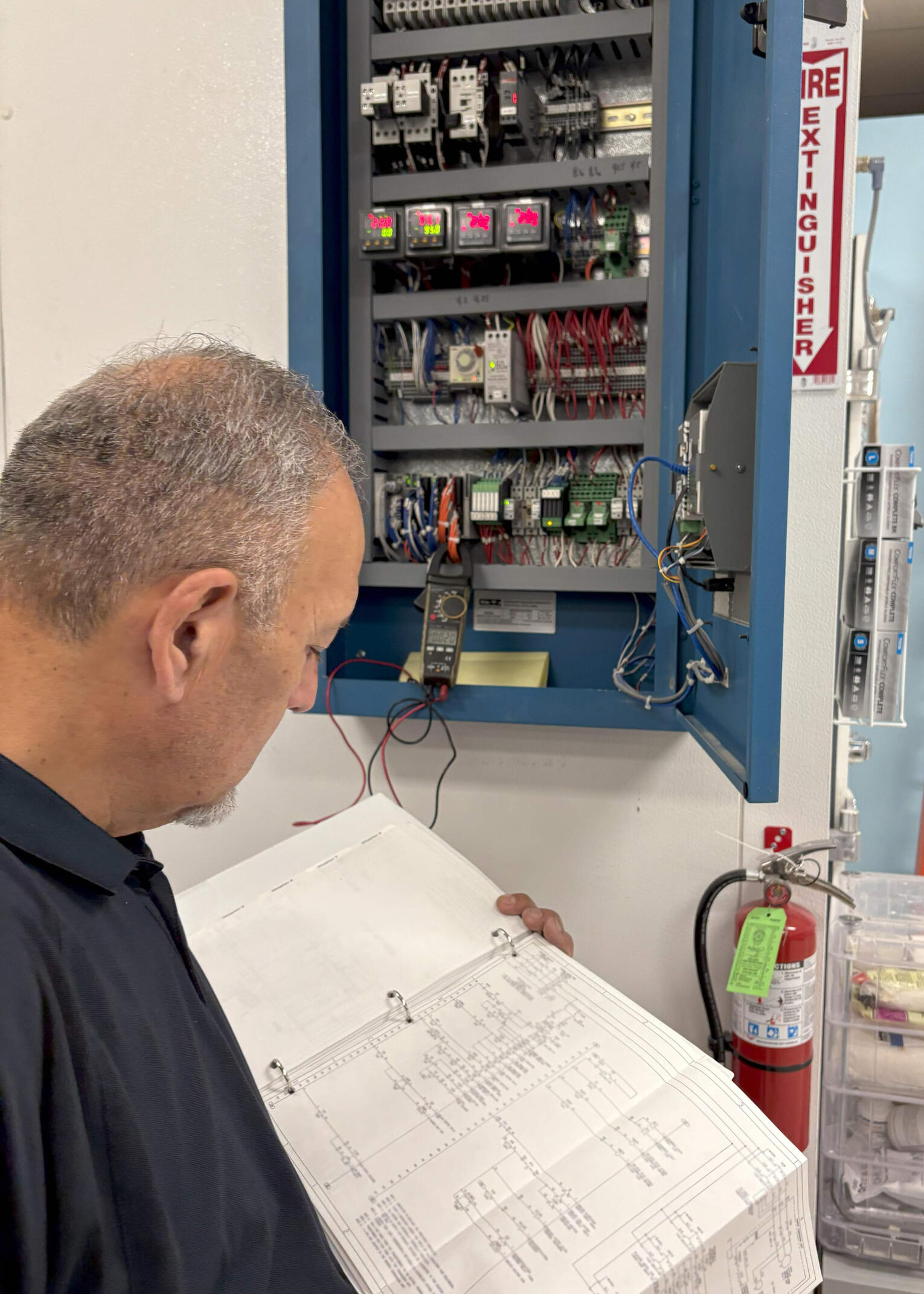 Technician Reviewing Electrical Schematics A technician examining detailed electrical schematics in front of an open control panel, filled with wiring and electrical components, in an industrial setting.